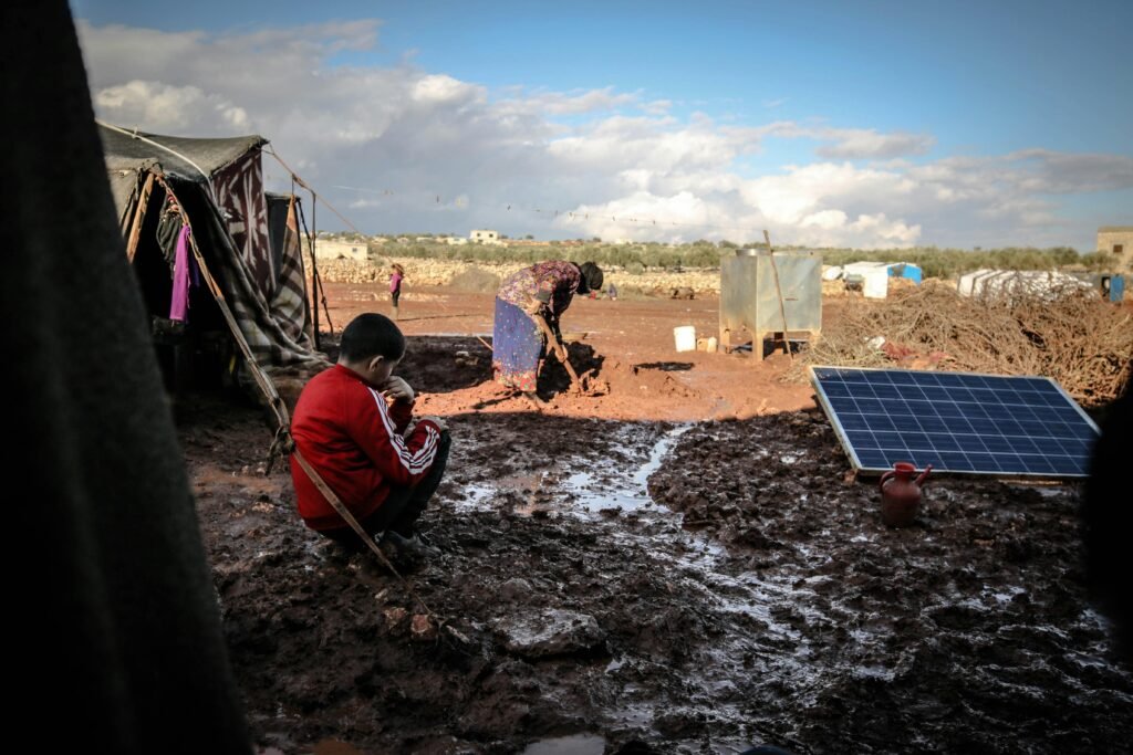 A boy and woman in a muddy refugee camp in Idlib, Syria with tents and solar panel.